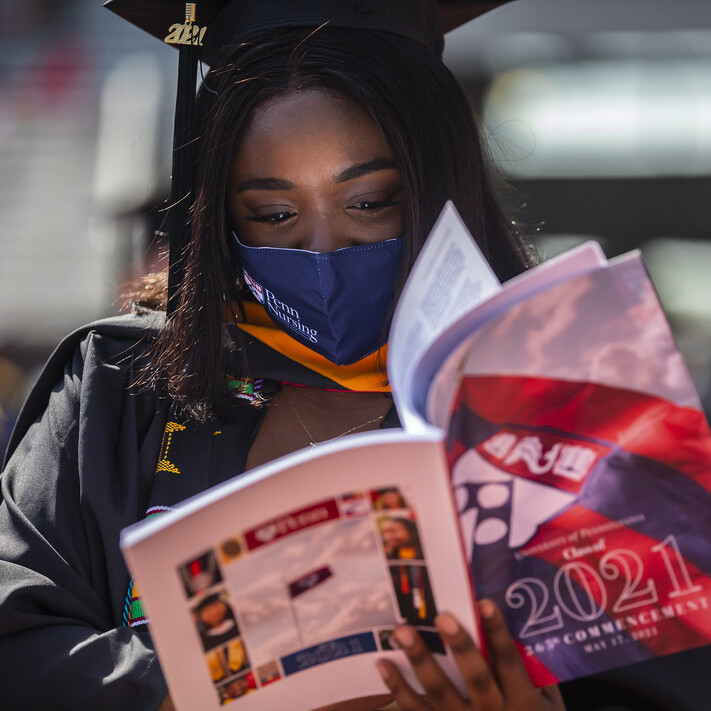 Graduating student in cap and gown reading the 2021 graduating class booklet at Franklin Field